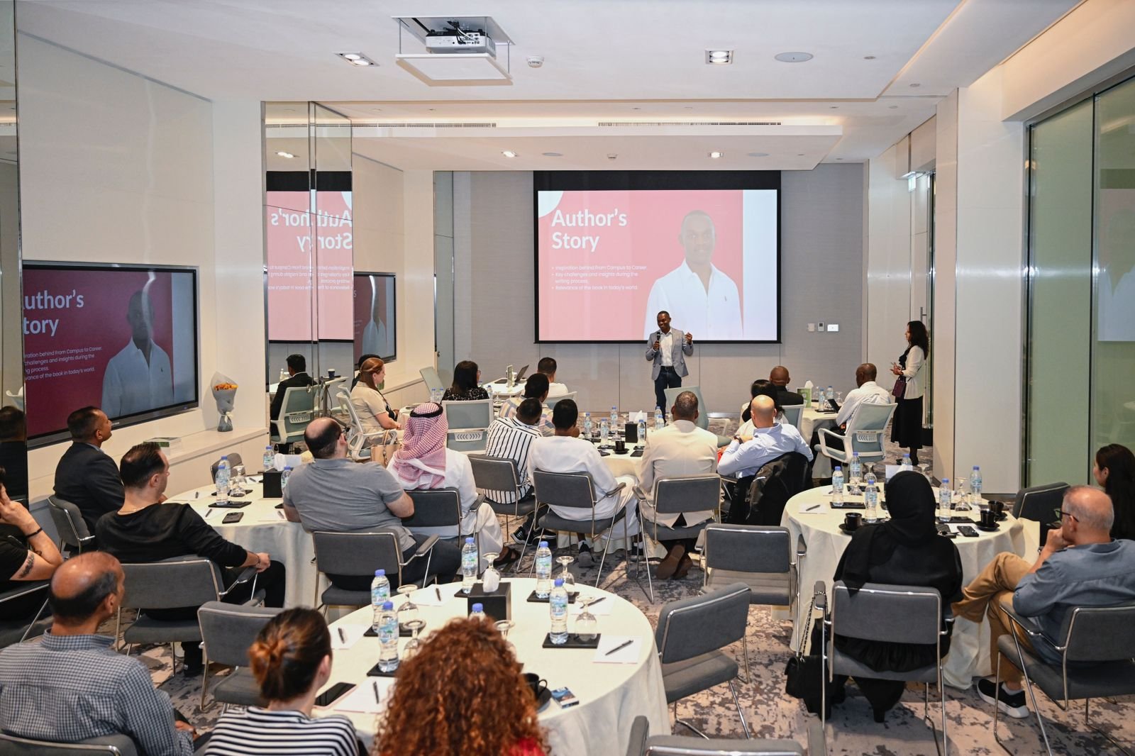 a group of people sitting around tables and a man standing in front of a screen
